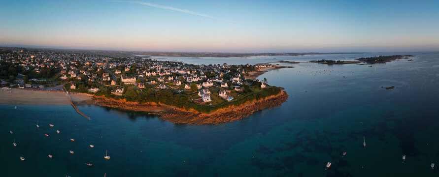 Aerial view of Plage du Kelenn where the warm glow of sunrise kisses the rocky coastline, contrasting with the cool, tranquil blue of the sea, Carantec, Bretagne, France.