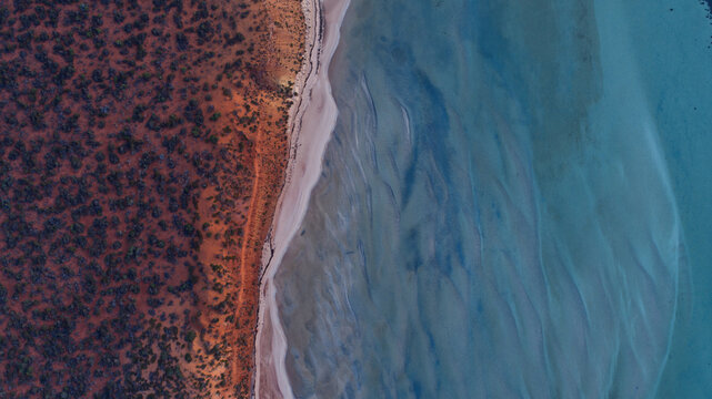 Aerial view of the vivid crimson earth meeting the tranquil, shallow turquoise waters of the coastline, revealing intricate sand patterns, Denham, Western Australia, Australia.