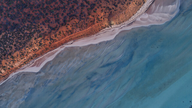 Aerial view of the vibrant red land meeting the clear turquoise water, separated by a thin sandy beach, Denham, Western Australia, Australia.