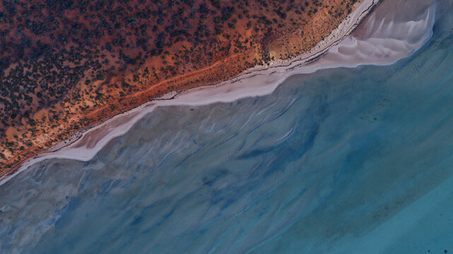 Aerial view of the stark contrast between the vibrant red earth and the pale sandy coastline meeting the tranquil blue-green ocean waters, Denham, Western Australia, Australia.