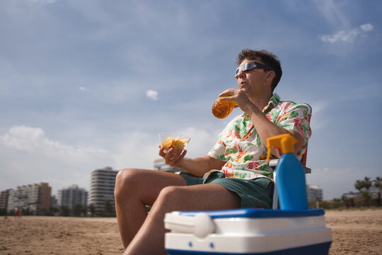 Man wearing eclipse viewing glasses, a colorful shirt, and shorts relaxing on a sandy beach, holding a bottle and eating chips while observing the sky