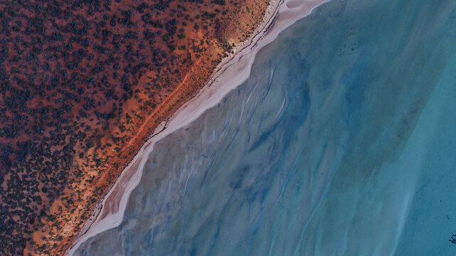 Aerial view of the reddish-brown land contrasting with a white sandy beach and the patterned blue ocean, Denham, Western Australia, Australia.
