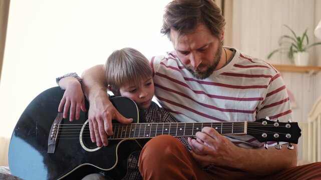 Handsome dad teaches his cute son to play the guitar sitting on sofa in the living room at home. Happy Father's Day. Father teaching his child to play guitar. Kid Learning music skill. Art education