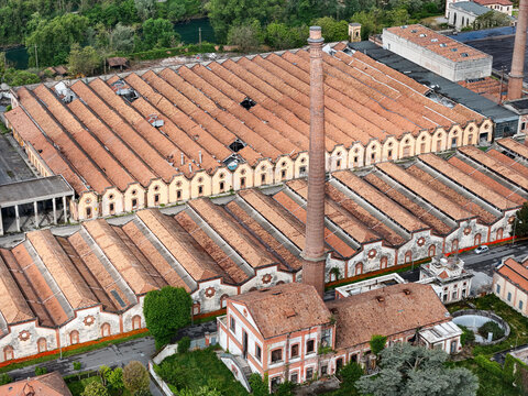 Aerial view of the Industrial Village of Crespi d'Adda featuring historic sawtooth factory roofs and a tall brick chimney stack surrounded by lush trees Capriate San Gervasio, Lombardia, Italy.