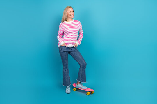 Smiling woman in pink striped sweater standing on a skateboard against a bright blue background, casual portrait of youth lifestyle and playful confidence