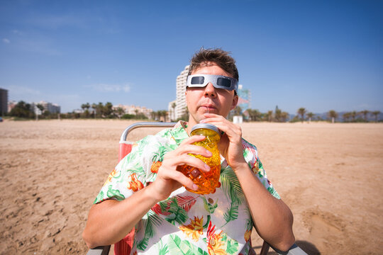Man in hawaiian shirt wearing solar eclipse glasses sits in a beach chair, sipping from a pineapple cup while watching the celestial spectacle under a clear blue sky