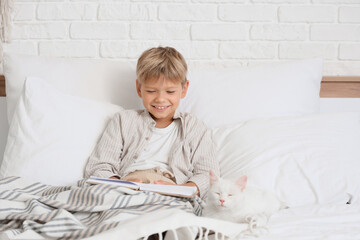 Naklejka premium Little boy with cat reading book under blanket in bedroom