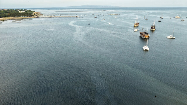 Aerial view of traditional phinisi boats and catamarans anchored in a calm tropical bay with a pier and distant island