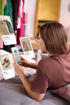Woman browsing clothes on smartphone with floating fashion interface in cozy room. Concept of online shopping, augmented reality fitting, ecommerce app and personalized outfit recommendations.