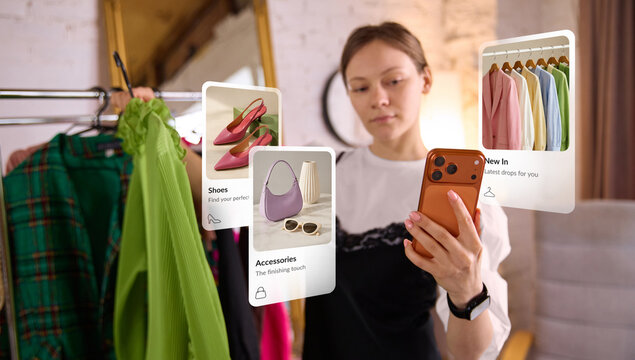Woman selecting clothes from rack while viewing digital fashion interface on smartphone. Concept of virtual styling, ecommerce technology, outfit selection and personalized shopping experience.
