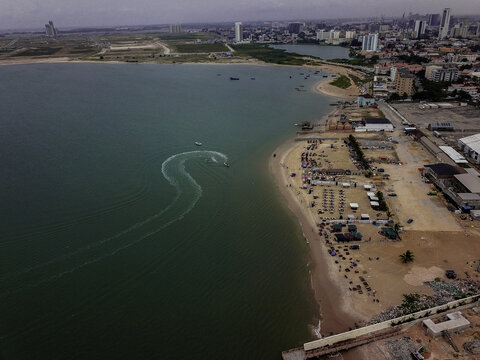 Aerial view of a jet ski carving S-shaped trails on the deep emerald waters beside a bustling sandy beach and an evolving urban skyline, Lagos, Lagos, Nigeria.