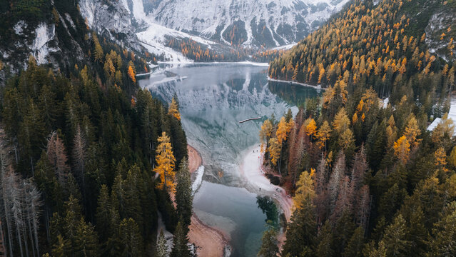 Aerial view of Lago di Braies, its calm surface mirroring snow-dusted peaks and a vibrant tapestry of autumn trees, Lago di Braies, Trentino-South Tyrol, Italy.