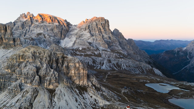 Aerial view of rugged peaks catching golden light, casting shadows over high alpine terrain and tranquil lakes, Tre Cime di Lavaredo, Veneto, Italy.