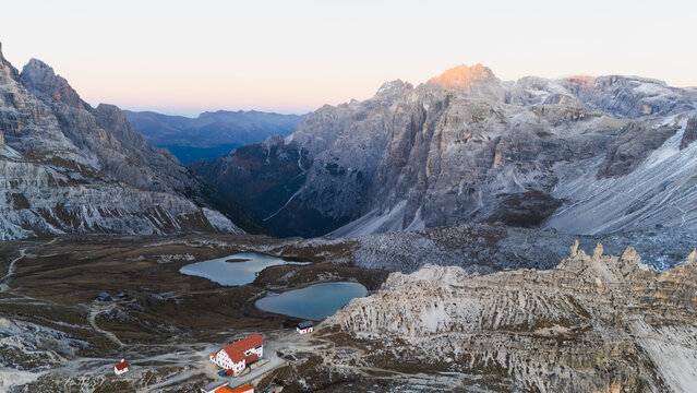 Aerial view of rugged, grey peaks and serene blue lakes contrasting with a warm-lit red-roofed refuge, Tre Cime di Lavaredo, Veneto, Italy.