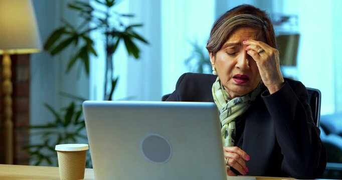 Senior Indian businesswoman in corporate attire having headache while working on laptop in modern cozy office space experienced female executive with white hair feeling stressed at her desk