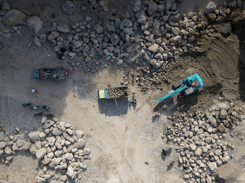 Aerial view of heavy machinery extracting and loading raw stone amidst vast grey rock piles and dusty earth, Klaten, Central Java, Indonesia.