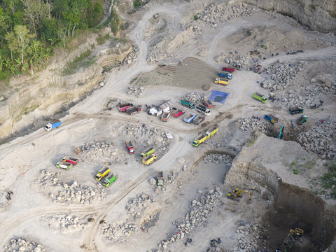 Aerial view of an active quarry with colorful trucks and excavators on pale, rocky terrain beside a green forest, Klaten, Central Java, Indonesia.