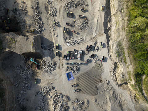 Aerial view of a vast open-pit quarry, with heavy machinery working the rugged grey terrain, bordered by a vibrant green forest, Klaten, Central Java, Indonesia.