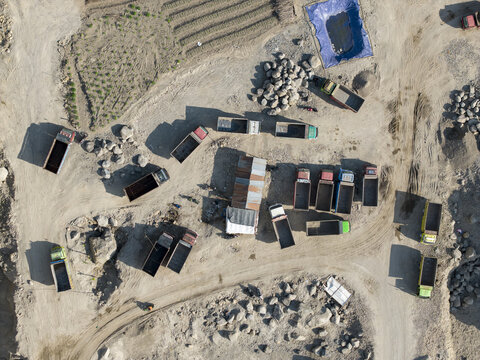 Aerial view of a dusty quarry, with dump trucks parked on rugged terrain next to scattered rock piles and a vibrant blue tarp, Klaten, Central Java, Indonesia.