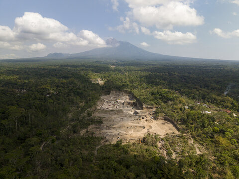 Aerial view of a vast, pale excavation site carved into the dense, emerald jungle, set against the dramatic silhouette of a majestic volcano with a smoky plume, Klaten, Central Java, Indonesia.