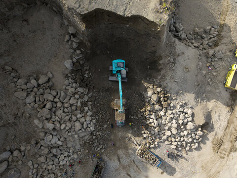 Aerial view of a teal excavator amidst a stark, rocky excavation site, contrasting textures of loose earth and heavy stones, Klaten, Central Java, Indonesia.