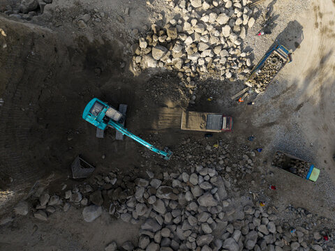 Aerial view of a vibrant teal excavator and dump trucks moving rugged grey rocks and sandy earth, Klaten, Central Java, Indonesia.
