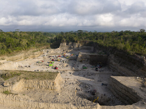 Aerial view of a vast quarry, where heavy machinery works amidst layers of earth and rock, contrasting with the surrounding lush greenery, Klaten, Central Java, Indonesia.
