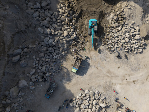 Aerial view of turquoise excavator stands out amid the grey rock and soil, loading trucks with earth in a bustling scene, Klaten, Central Java, Indonesia.