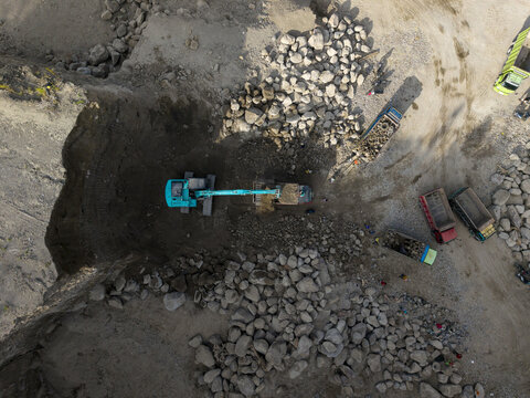 Aerial view of a vibrant turquoise excavator digging a deep, dark pit amidst rugged piles of grey rocks and waiting dump trucks under the bright sun, Klaten, Central Java, Indonesia.