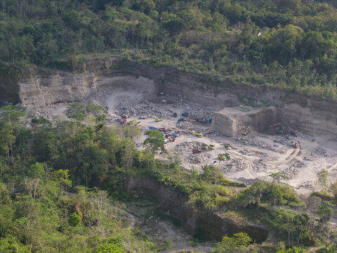 Aerial view of a vast quarry operation, where the scarred earth and raw textures of excavation starkly contrast with the surrounding lush green canopy, Klaten, Central Java, Indonesia.