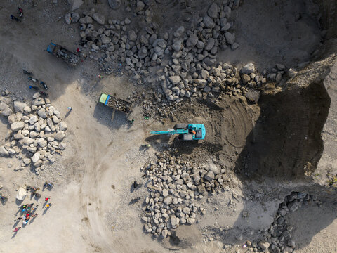 Aerial view of a bustling open-pit quarry with a vibrant turquoise excavator digging into the earth amidst vast piles of grey rocks, Klaten, Central Java, Indonesia.