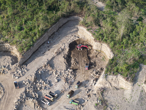 Aerial view of a stark contrast where verdant tropical forest meets a raw, excavated earth pit, revealing heavy machinery and trucks at work, Klaten, Central Java, Indonesia.