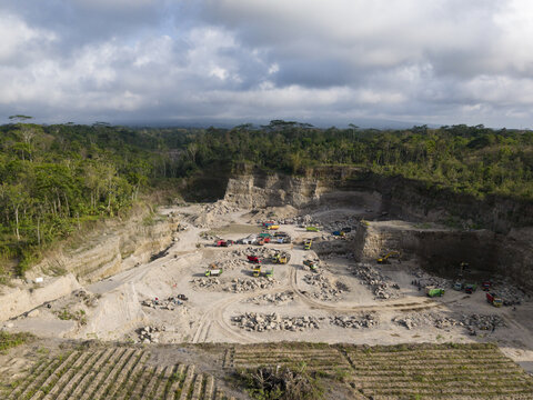 Aerial view of a vast open-pit mine carving into dense green tropical rainforest, revealing stark light-toned earth and heavy machinery at work, Klaten, Central Java, Indonesia.