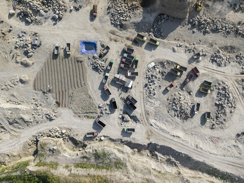 Aerial view of a vast, sun-drenched quarry, where earth-toned trucks navigate rugged terrain amidst stark piles of rock and gravel, Klaten, Central Java, Indonesia.