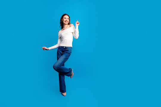 Smiling young woman in white off-shoulder sweater and jeans dancing playfully against bright blue studio background, casual fashion portrait conveying joy, confidence and carefree energy