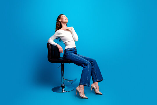 Happy young woman posing on a modern stool against a bright blue background in casual jeans and heels, stylish portrait conveying confidence, relaxation and contemporary fashion