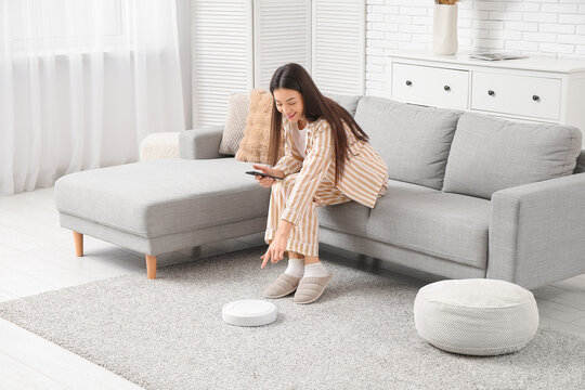 Young woman in pajamas using mobile phone on sofa with robot vacuum cleaner at home