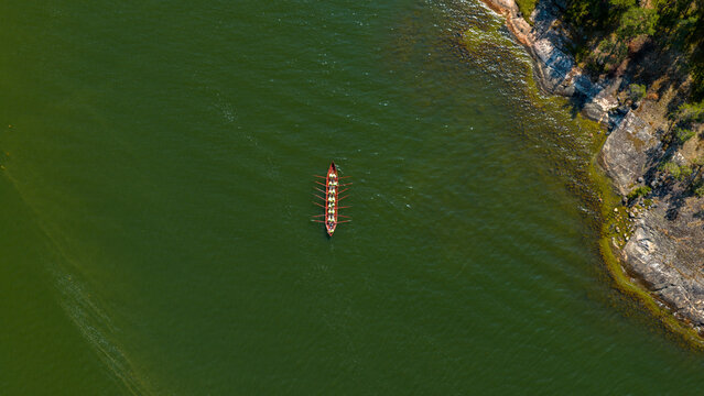 Aerial view of a long rowing boat gliding across emerald waters beside rugged, tree-lined granite shores, Hiittinen, Southwest Finland, Finland.