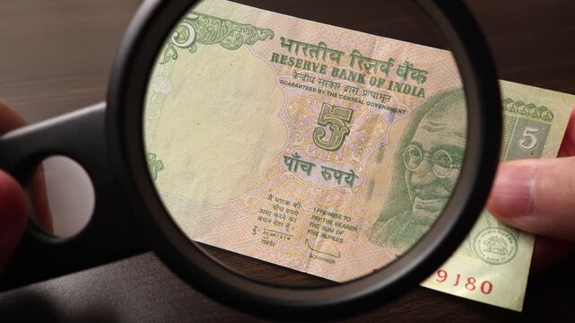Examination of 5 Indian rupees (INR) cash money with magnfication glass, loupe. Concept of authenticity check, fight against counterfeiting of banknotes, anti fraud measures in India. Close up view.