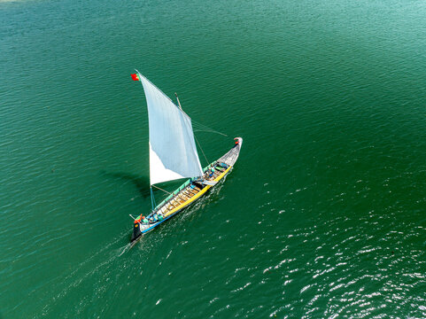 Aerial view of a traditional sailing boat with a stark white sail gliding across the deep emerald waters, leaving a subtle wake, Torreira, Aveiro District, Portugal.