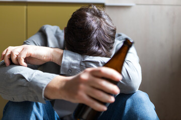 Young man holding beer bottle with head down indicating alcohol misuse and emotional distress for...
