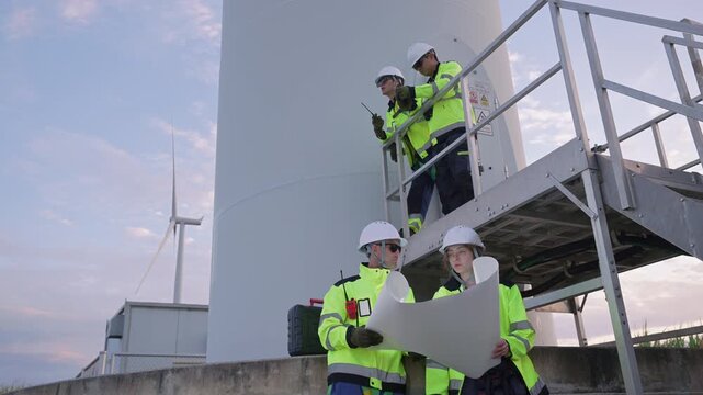 Engineers wearing safety helmets and jackets inspecting a wind turbine at wind farm at sunset. Concept renewable energy, sustainable technology wind power maintenance and environmental engineering.