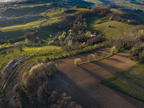 Aerial view of sun-drenched rolling hills contrasting with deep shadows across freshly tilled earth and vibrant early spring foliage, Tortona, Piedmont, Italy.