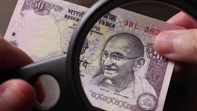 Examination of 50 Indian rupees (INR) cash money with magnfication glass, loupe. Concept of authenticity check, fight against counterfeiting of banknotes, anti fraud measures in India. Close up view.