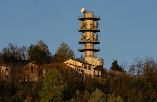 Aerial view of a communication tower and buildings on a wooded hill under a bright sky, Castellania, Piedmont, Italy.