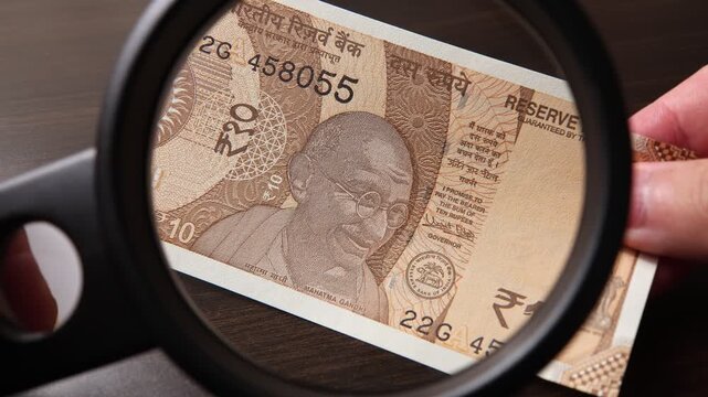Examination of 10 Indian rupees (INR) cash money with magnfication glass, loupe. Concept of authenticity check, fight against counterfeiting of banknotes, anti fraud measures in India. Close up view.