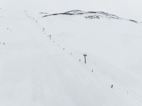 Aerial view of white ski slopes with lift lines guiding tiny figures towards the stark, dark volcanic rock, Piano Provenzana, Sicilia, Italy.
