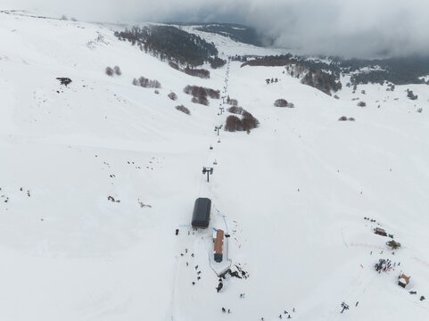 Aerial view of a vast white blanket of snow covering the mountain slopes, punctuated by dark ski lift structures and scattered trees, under a muted sky, Piano Provenzana, Sicilia, Italy.