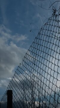 Vertical video of a metal chain link fence silhouette against a deep blue sky with moving white clouds in 4K.