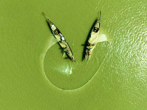 Aerial view of two slender fishing boats casting a wide net across the calm, emerald waters, Bogura, Rajshahi Division, Bangladesh.